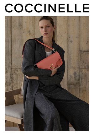 Bolso de piel rosa con forma curva, acentos de hardware dorado y una correa ajustable, sostenido contra un modelo que lleva un traje gris.