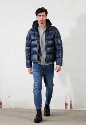 Giacca imbottita blu navy con cappuccio nero, maglione grigio e jeans blu. Presenta una finitura lucida e polsini a costine. Stivali neri con lacci.