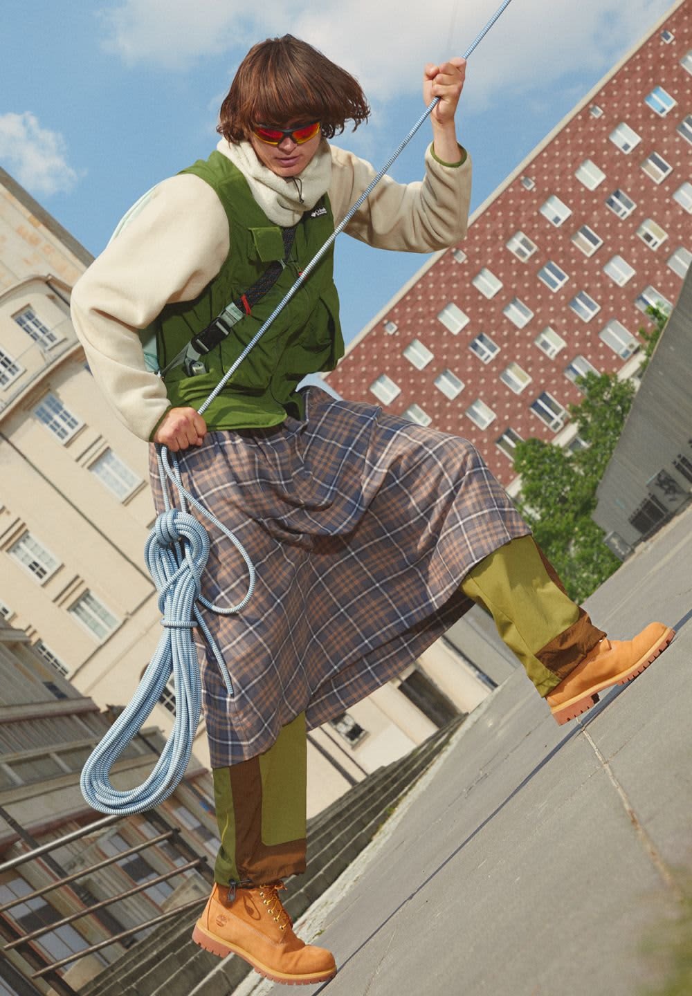 Green vest with black accents over a cream jumper, checked skirt, olive trousers, and orange boots. Holding climbing rope, outdoor setting.