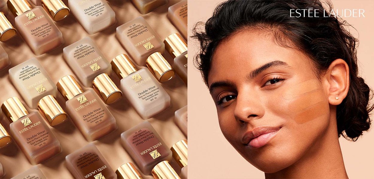 File di bottiglie di fondotinta Estée Lauder Double Wear accanto a una donna con campioni di fondotinta sulla guancia.