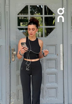 Young woman in black pinstripe two-piece outfit stands by gray door, holding phone and tangled earphones, with trees reflected in window behind.