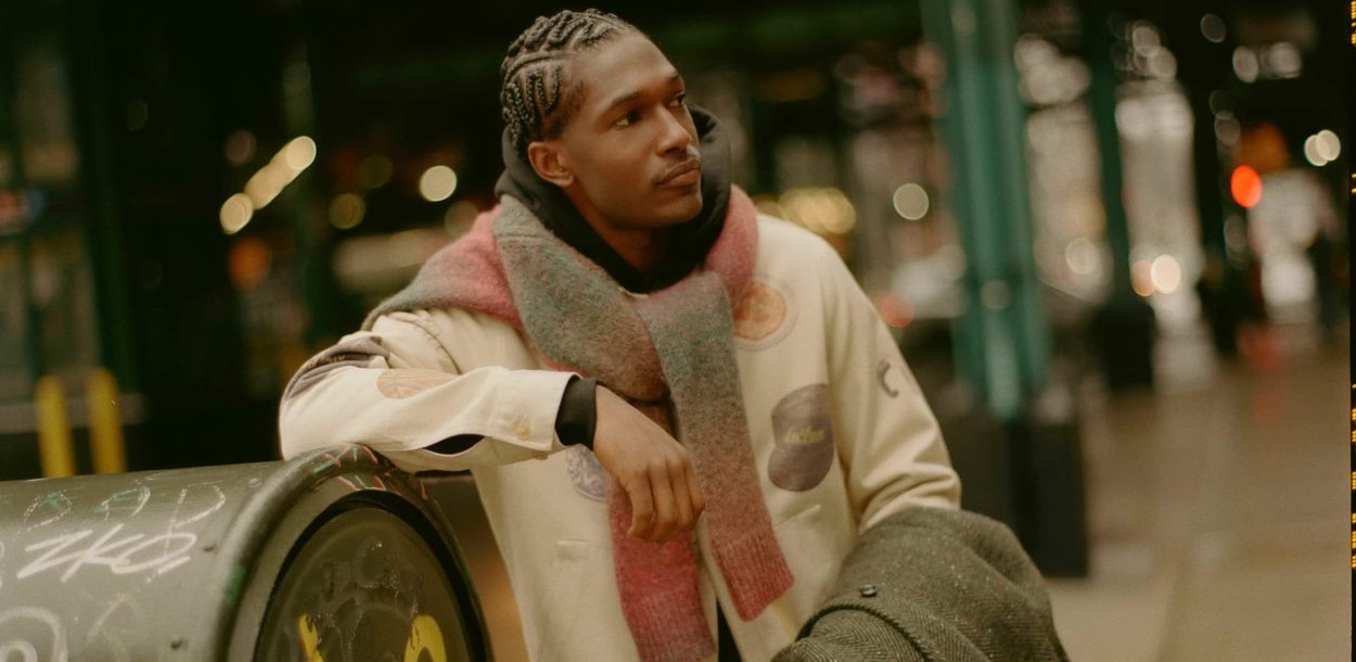 Ung mand med fletninger iført halstørklæde og let jakke læner sig op ad en graffiti-beklædt postkasse om natten i en bymæssig setting.