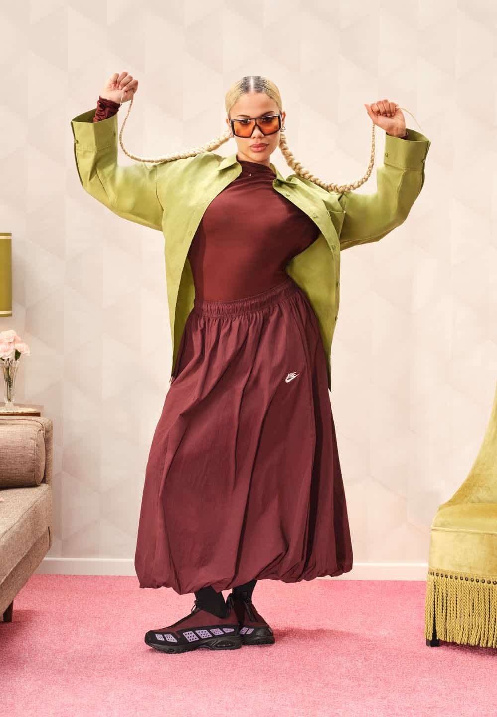 Camicia verde oversize, top aderente bordeaux, gonna bordeaux fluida con logo Nike e scarpe da ginnastica nere. Capelli intrecciati e occhiali da sole.