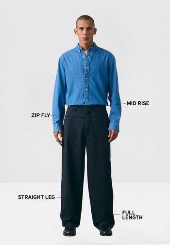 Hombre de pie vistiendo una camisa azul abotonada y pantalones negros de pierna recta, de longitud completa, con detalles de tiro medio y cierre de cremallera etiquetados.