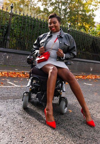 Svart läderjacka, grå klänning, röd handväska och röda högklackade skor. Rullstol med grå sits, utomhusmiljö med höstlöv och ett staket.