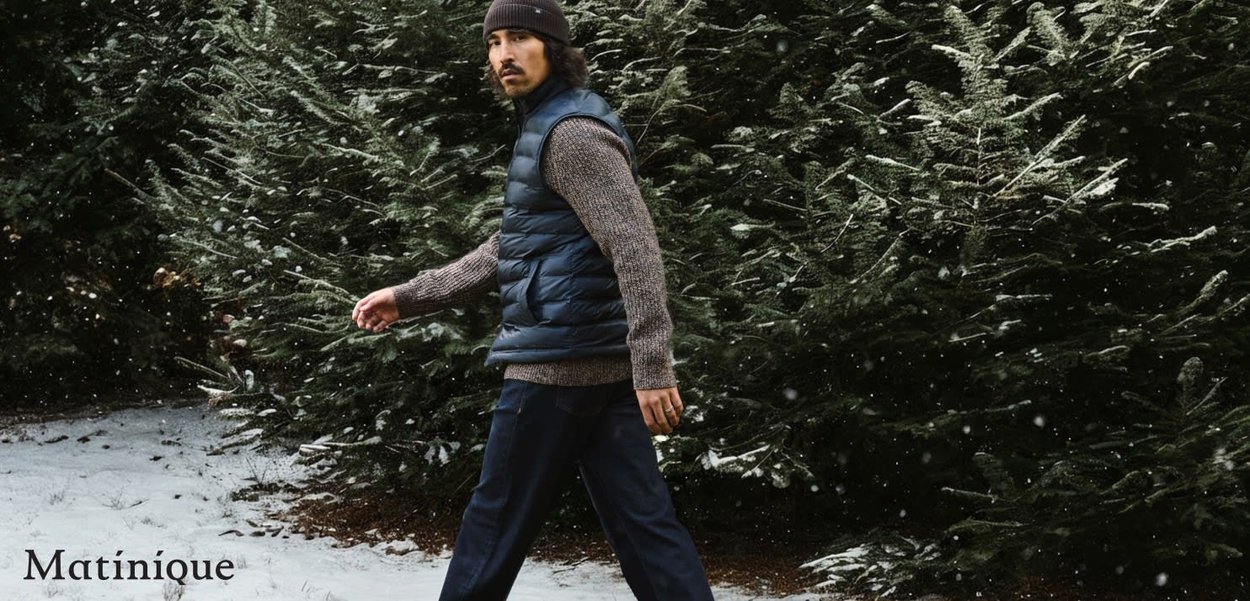 Gepolsterte marineblaue Weste über einem strukturierten braunen Pullover, kombiniert mit dunkler Hose. Schnee bedeckt den Boden in einer bewaldeten Umgebung.