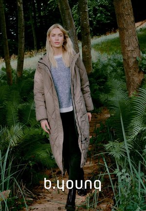 Lungo cappotto imbottito beige con cappuccio, caratterizzato da un design trapuntato e una zip centrale. Abbinato a un maglione a righe blu e pantaloni neri.
