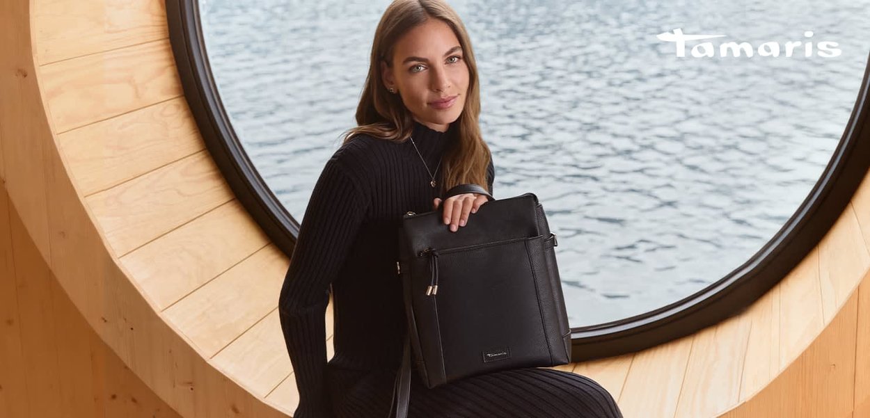 Schwarze Ledertasche mit strukturierter Oberfläche und einem oberen Griff, ausgestattet mit einer Reißverschlusstasche auf der Vorderseite. Vor einem runden Fenster platziert.