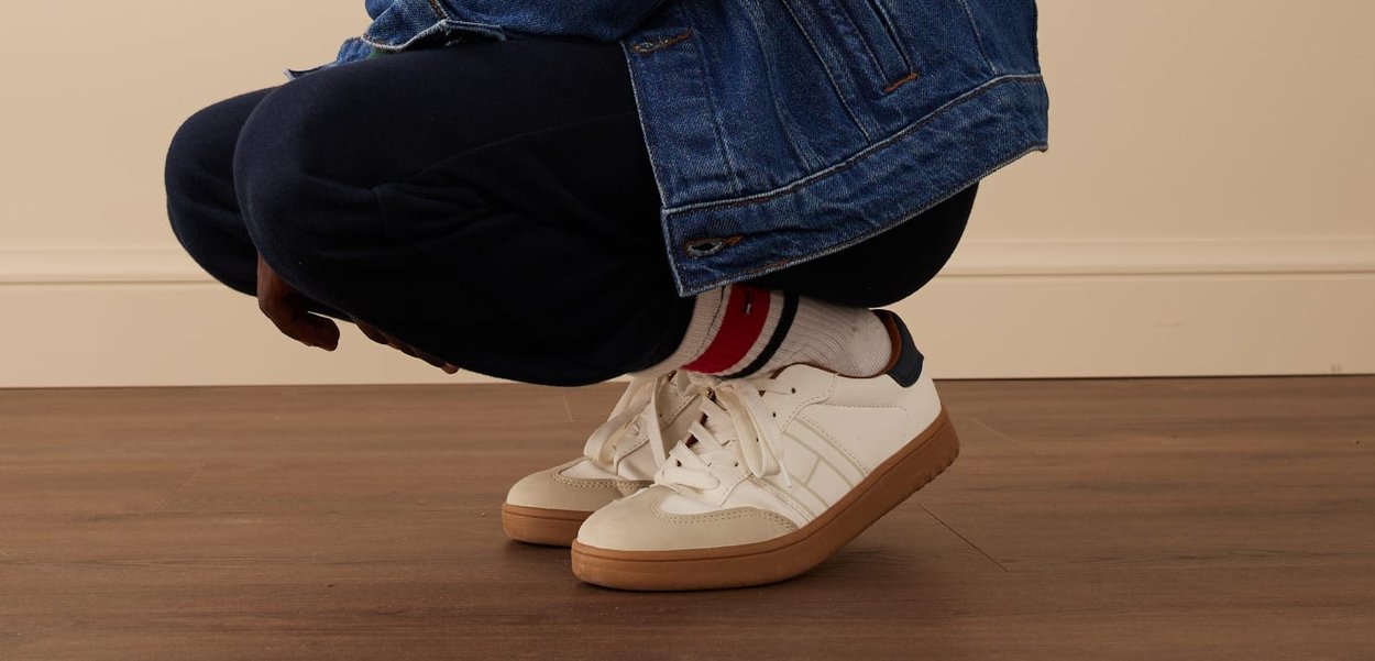 Persona in ginocchio dentro casa, che indossa scarpe da ginnastica bianche con suole color gomma, calze bianche con strisce rosse e nere, pantaloni scuri e una giacca di jeans.
