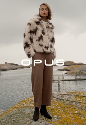Woman stands on rocky shore wearing spotted faux fur jacket, wide brown pants, and black boots under cloudy sky.