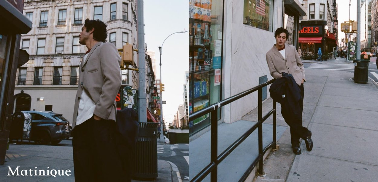 Giovane uomo con giacca beige in piedi e appoggiato a una ringhiera nera sul marciapiede della città, con edifici alti e segnali stradali intorno.