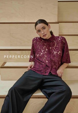 Chemise en dentelle bourgogne à manches courtes, avec un col et des motifs floraux, associée à un pantalon noir ample. Escalier en bois en arrière-plan.