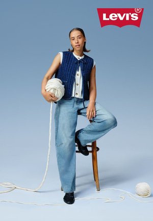 Marineblauwe gewatteerde vest over een witte top, gecombineerd met lichtblauwe wijde jeans. Model houdt witte garen vast en zit op een houten kruk tegen een blauwe achtergrond.