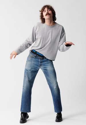 Graues Langarmshirt, ausgewaschene blaue Denim-Jeans mit Used-Details und schwarze Schuhe. Sichtbarer blauer Bund mit dem Schriftzug "HAPPY".
