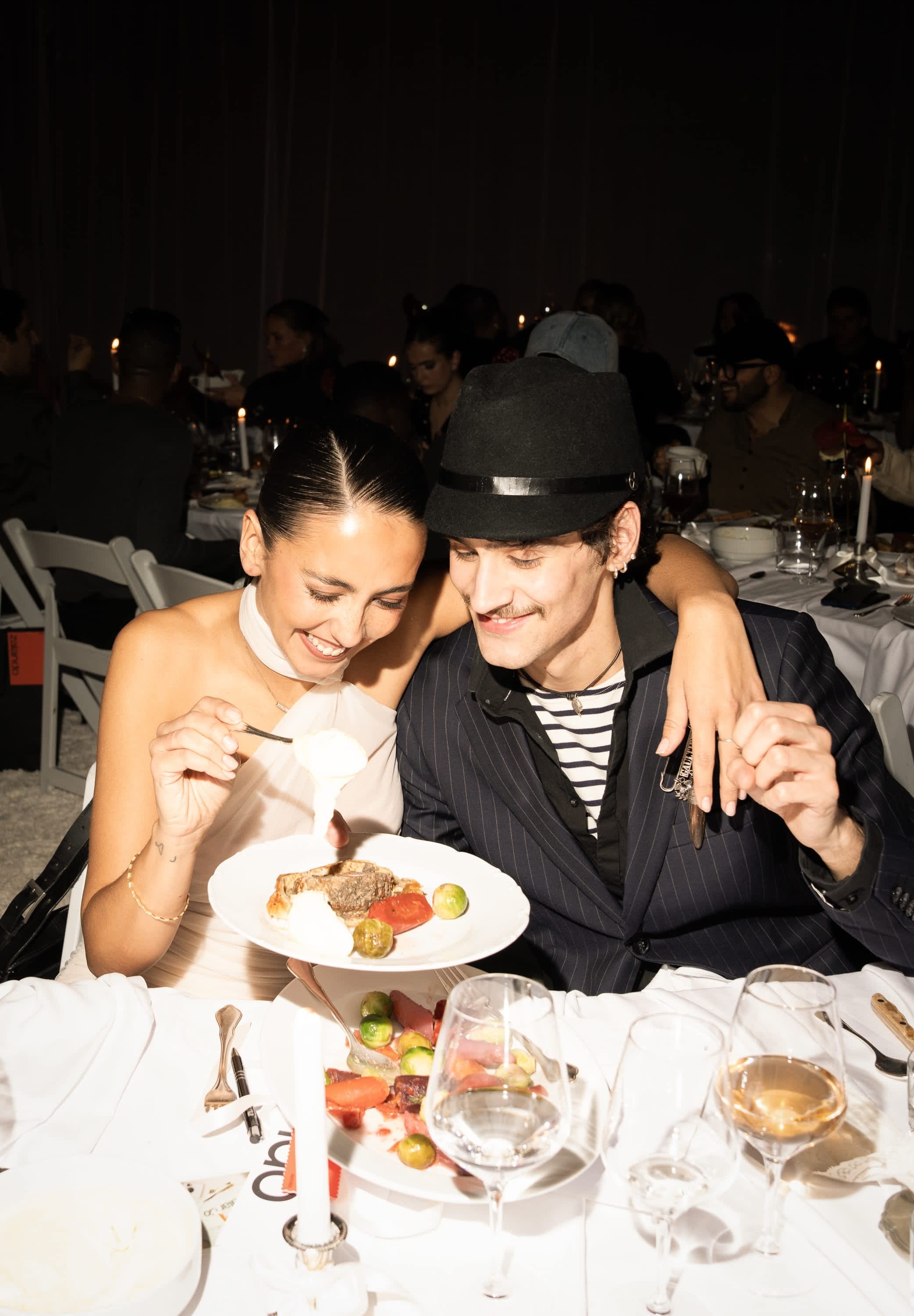 Två personer delar en måltid vid ett dukat bord. Kvinnan håller en tallrik med kött och grönsaker och bär en ljus klänning. Mannen bär en randig skjorta och hatt.