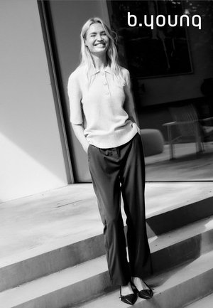 Hellgraues, kurzärmeliges Strickoberteil mit Knopfdetail, kombiniert mit schwarzen, weit geschnittenen Hosen und spitzen, schwarzen Ballerinas, stehend im Freien.