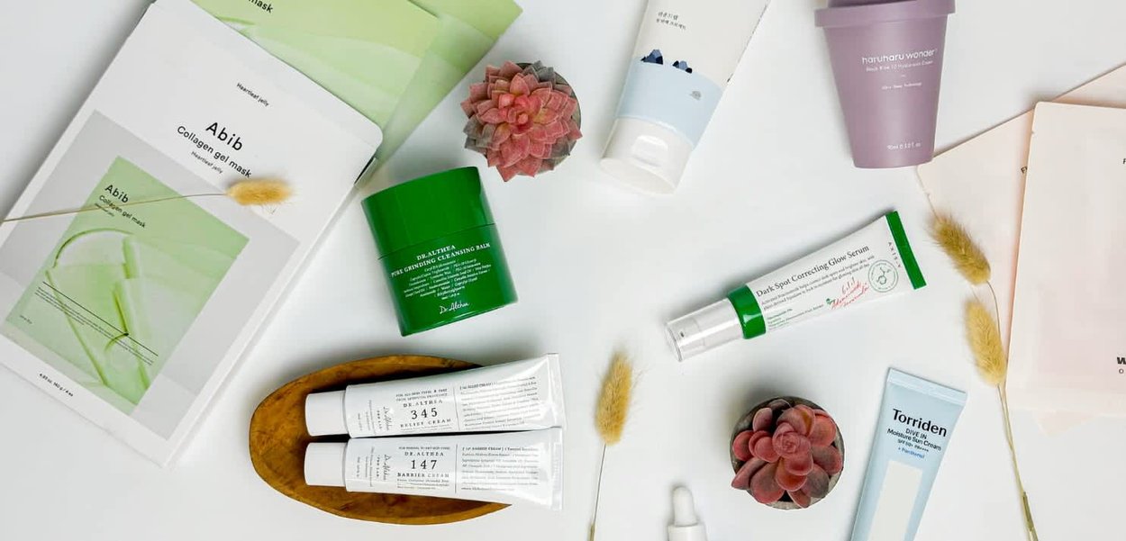 Collection de produits de soin de la peau : baume nettoyant vert, crèmes blanches, hydratant violet et masque en gel, disposés sur un fond clair avec des plantes séchées.