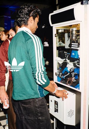 Green athletic jacket with white stripes, dark pants, and a vending machine displaying black and blue socks. White hardware on machine.