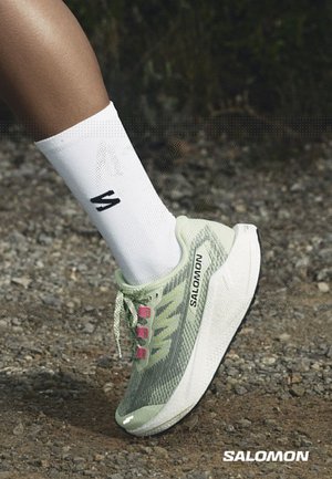 Fod iført en lysegrøn Salomon løbesko med pink snørehuller og en hvid sok med sort logo på en grusoverflade.