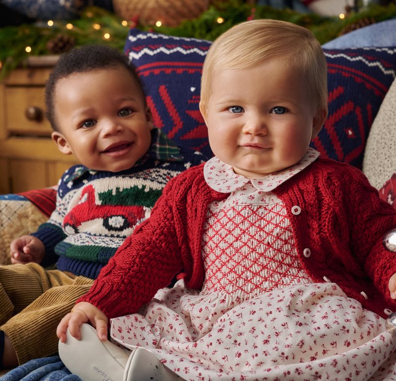 Deux enfants sont assis ensemble, l'un portant un pull en tricot vert avec un motif de voiture rouge, l'autre portant un cardigan rouge sur une robe blanche à fleurs.