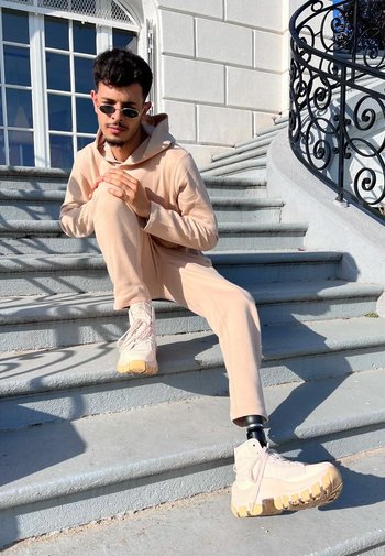 Beige huvtröja och joggers-outfit med texturerad tyg. Klumpiga beige och vita sneakers, som visar en protesben, sittande på stensteg.