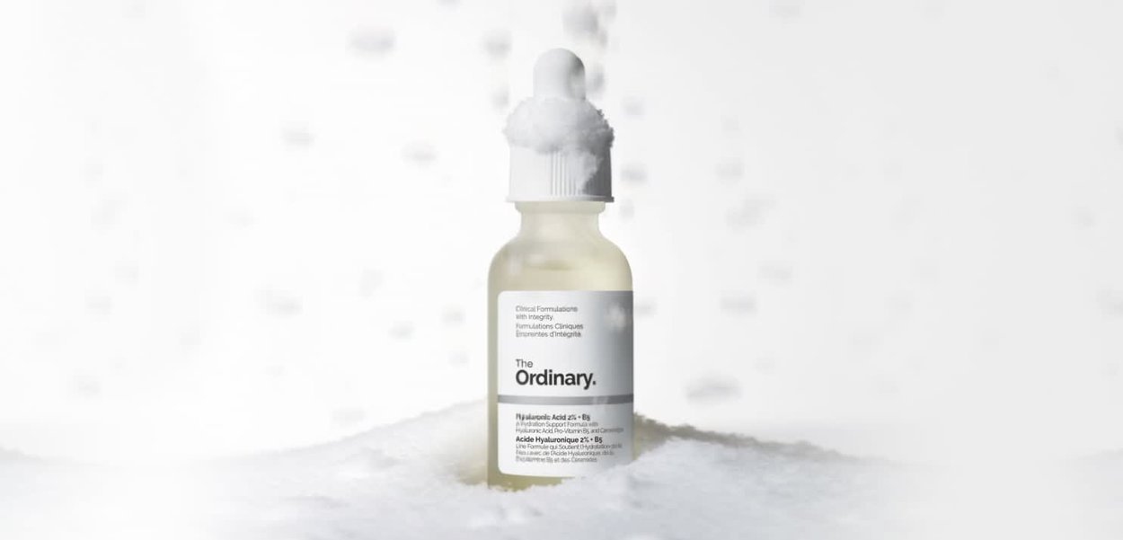 Heldere glazen druppelfles met witte dop, gevuld met een lichtgeel serum, gelabeld met "The Ordinary" in zwarte tekst, omgeven door zachte witte deeltjes.