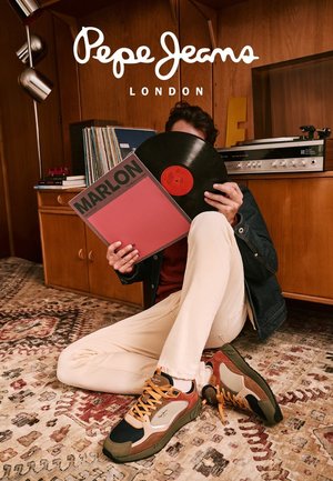 Persona sentada en una alfombra con un diseño estampado, sosteniendo un disco de vinilo y su funda, junto a un mueble de madera con un tocadiscos y discos apilados.