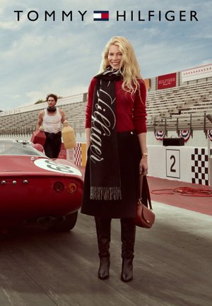 Model in a red sweater, black skirt, and black scarf with "Hilfiger" text, standing near a red race car with visible sponsor graphics.