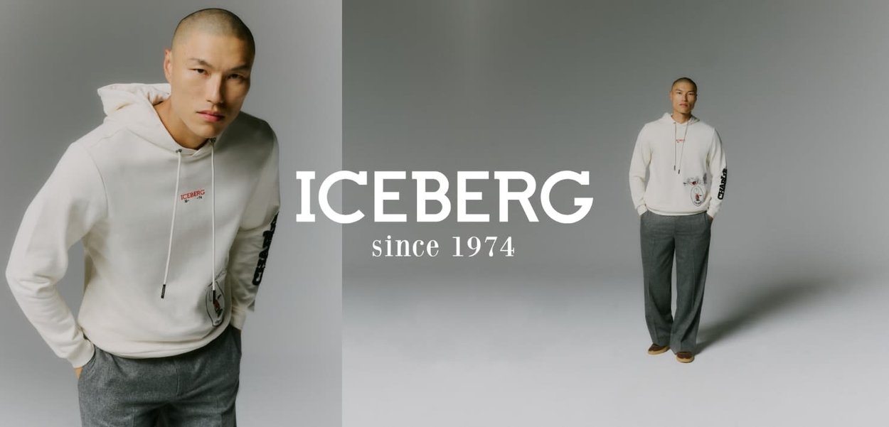 Weißer Kapuzenpullover mit Kordelzug, mit einem grafischen Druck und rotem Logo. Graue strukturierte Hose, kombiniert mit braunem Schuhwerk. Neutrale Hintergrund.