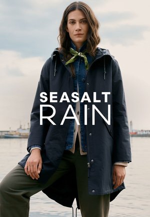 Vrouw in een marineblauwe regenjas, denim jasje, groene sjaal om haar hals en olijfgroene broek, staand bij een rustige waterkant onder een bewolkte lucht.