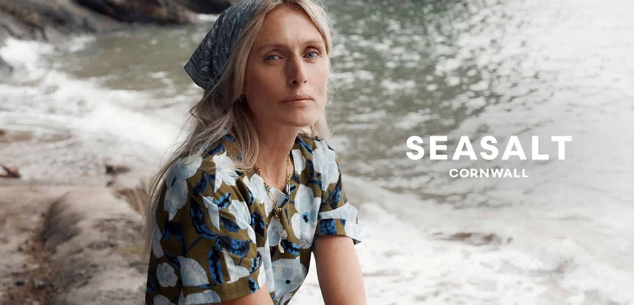 Lyshåret kvinde med et blåt hovedtørklæde med mønster og blomstret skjorte, der sidder på en sten ved havet med bølger bag sig.