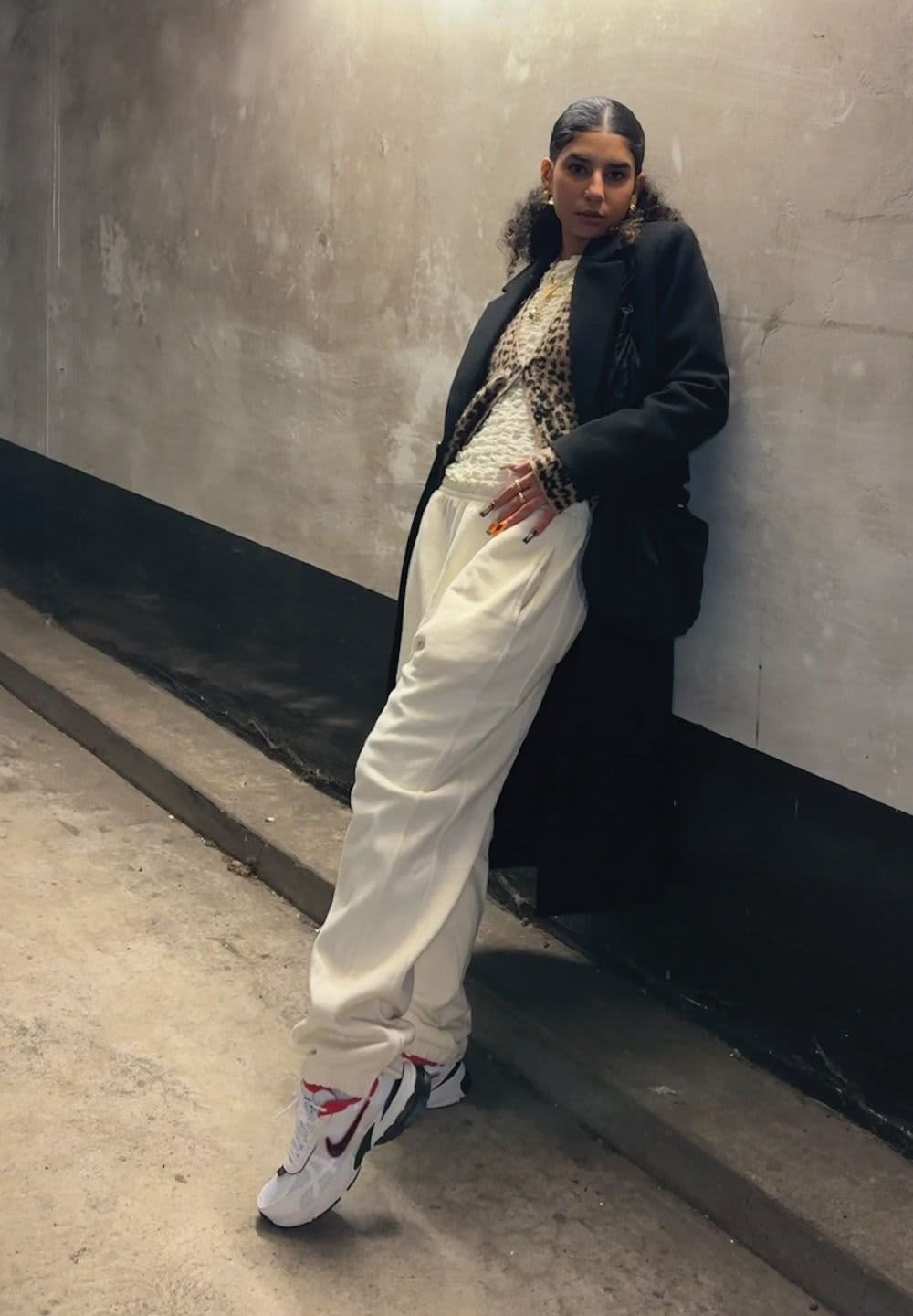 Black coat, leopard print sweater, white baggy trousers, and white sneakers with red and black accents. Smooth textures and layered look.