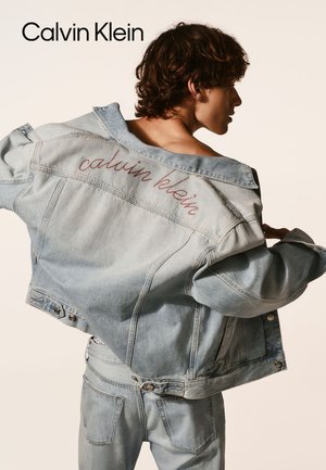 Light blue denim jacket with a relaxed fit, visible stitching, and "calvin klein" embroidered in red on the back. Textured collar and buttons.