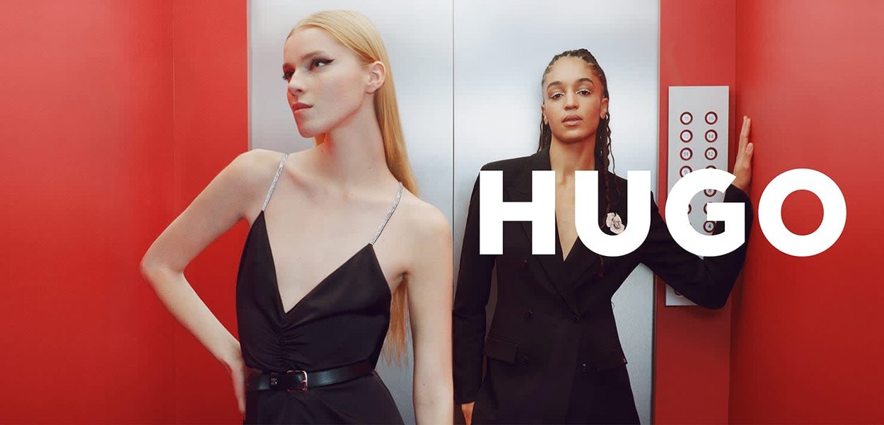 Deux femmes dans un ascenseur : l'une porte une robe noire avec des accents argentés, l'autre un blazer noir orné d'une broche florale. Fond rouge.