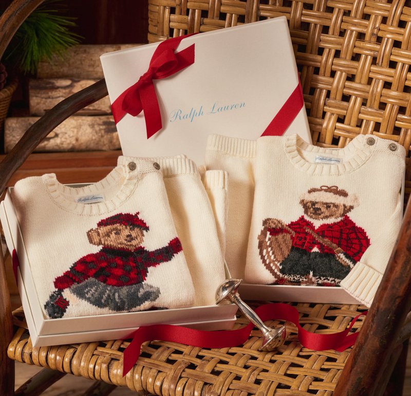Zwei cremefarbene Pullover in einer Geschenkbox mit bestickten Bären in karierten Outfits. Ein Bär hält eine Angelrute, der andere läuft.