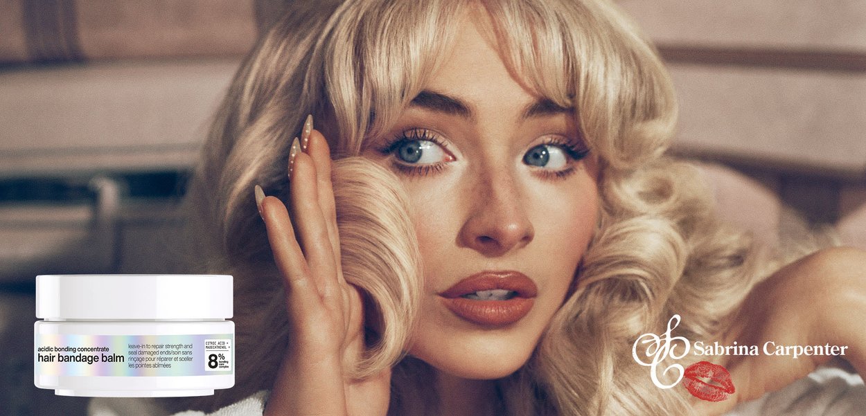 Blondes Frau mit gestylten Locken berührt ihr Haar und präsentiert einen Behälter mit Haarwundbalsam; das "Sabrina Carpenter"-Logo mit rotem Lippenstift-Abdruck ist sichtbar.