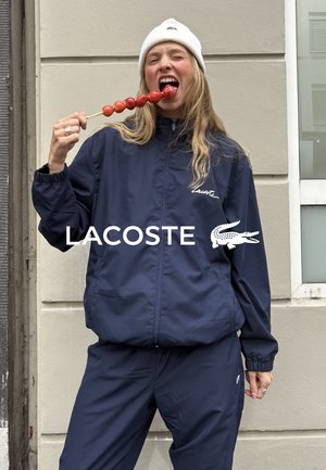 Veste coupe-vent bleu marine avec pantalon assorti, dotée d'une fermeture éclair à l'avant, de poignets élastiques et d'un logo sur la poitrine et la manche. Tient des aliments rouges sur un bâton.