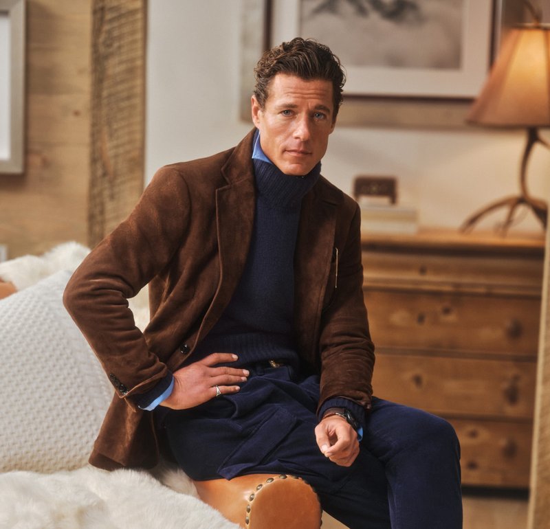 Uomo con giacca di camoscio marrone e dolcevita blu scuro seduto sul bracciolo di una poltrona in un soggiorno accogliente e illuminato dolcemente.