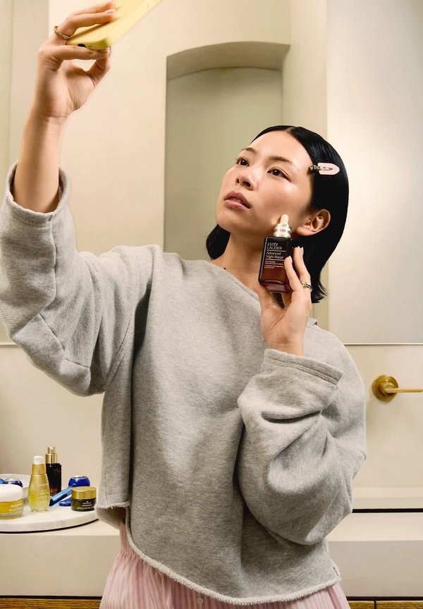 Vrouw brengt Estée Lauder Advanced Night Repair serum aan op haar gezicht terwijl ze een gele telefoon vasthoudt in een badkameromgeving.