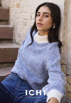 Blue and white striped sweater with a ribbed texture and a white collar, paired with dark blue high-waisted jeans, against a stone wall.