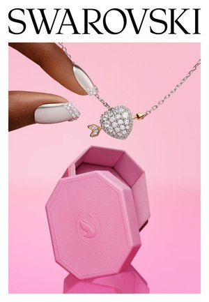 Hand houdt een zilveren hartvormige hanger met kristallen en een gouden pijl aan een zilveren ketting boven een open, roze, achthoekig sieradendoosje.