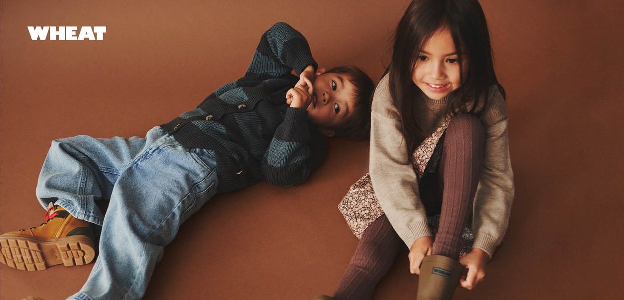 Two children lie on a brown surface. One wears a green striped sweater and blue jeans, the other a floral dress with ribbed brown tights and boots.