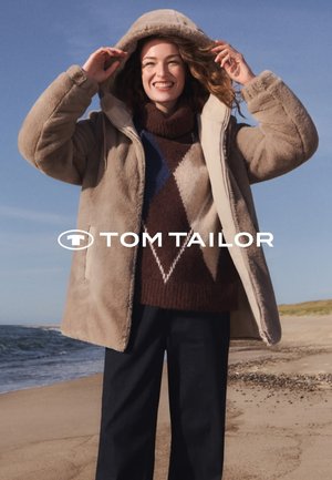 Beiger Fleece-Mantel mit Kapuze, kombiniert mit einem braunen Strickpullover mit Rautenmuster und dunklen Hosen. Küstenlandschaft im Hintergrund.