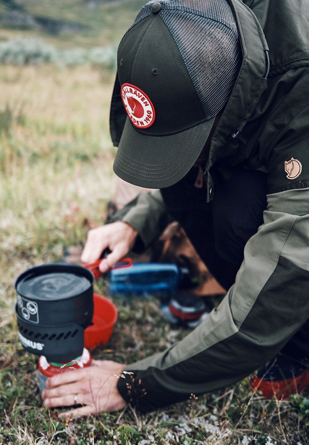 Campingkök med svart kastrull, röd skål och blå vattenflaska på gräset. Person i en khakifärgad jacka och grön keps förbereder mat utomhus.
