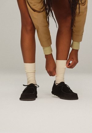 Persona che indossa pantaloni beige e calzini color crema si sistema il calzino su una gamba, scarpe nere con lacci su sfondo bianco.