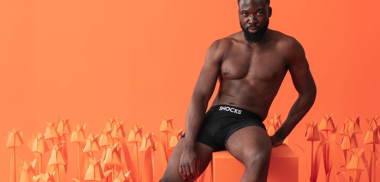 Oberkörperfreier Mann mit Bart, der schwarze Snocks-Unterwäsche trägt, sitzt auf einem orangefarbenen Block zwischen stilisierten orangen Papier-Tulpen vor orangefarbenem Hintergrund.