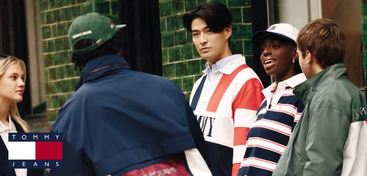 Cinque giovani adulti di diverse etnie stanno chiacchierando all'aperto, vestiti con abiti casual a righe e a tinta unita, con uno sfondo di muro piastrellato verde.