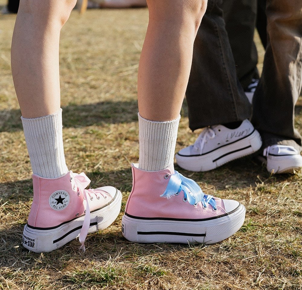Pink high-top sneakers with a platform sole, blue laces, and white rubber toe cap. Worn with ribbed white socks on grassy ground.