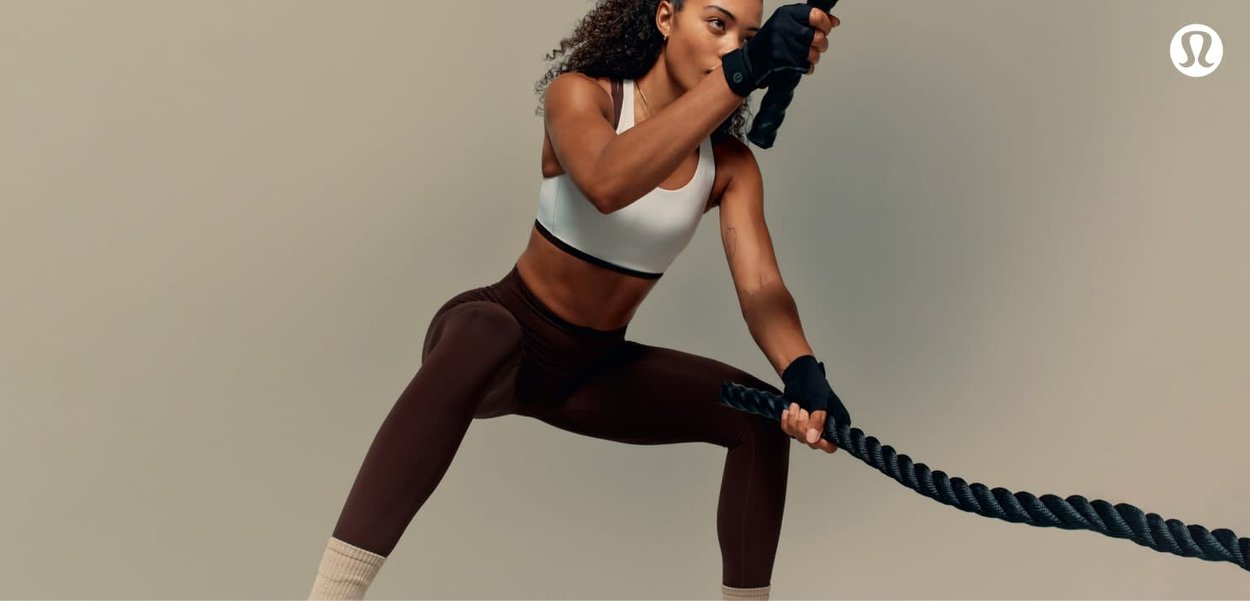 Nainen treenivaatteissa, joka pitää ja vetää paksua harjoitusköyttä dynaamisessa, matalassa kyykkyasennossa yksinkertaista taustaa vasten.