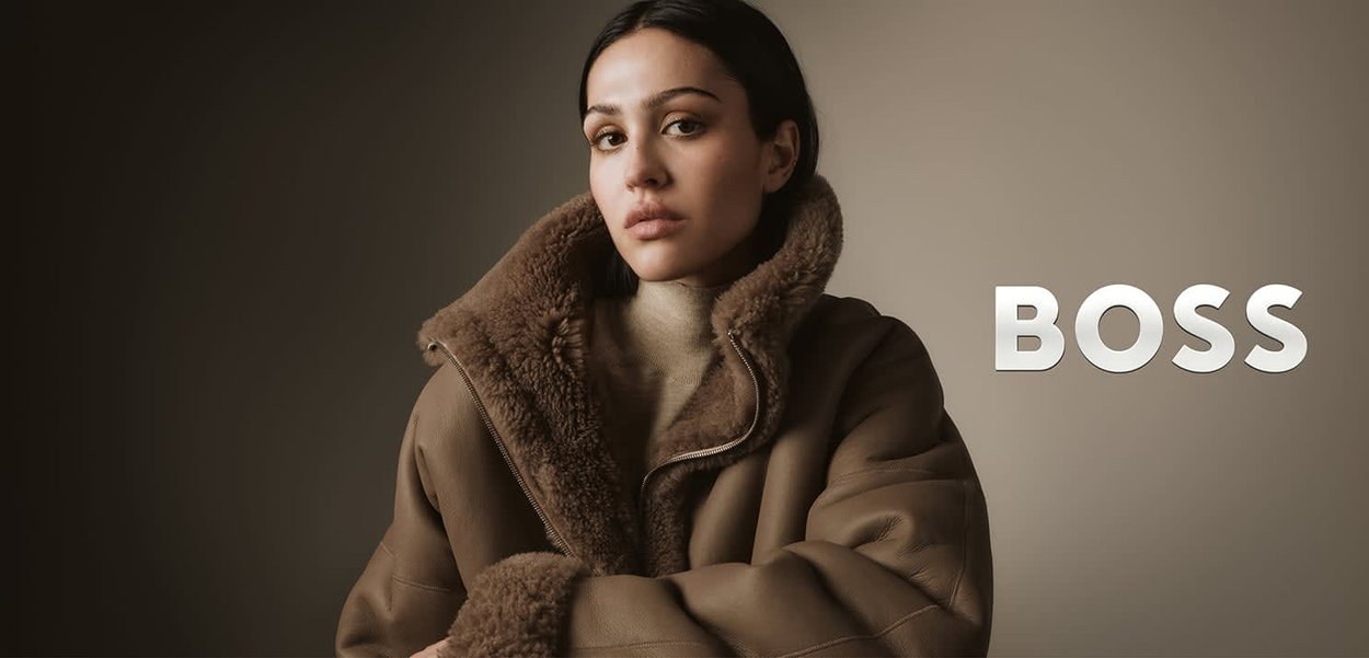 Bruine shearling jas met een brede kraag en ritssluiting, gedragen over een lichtbeige trui, met een zachte textuur. Het merk is "BOSS."